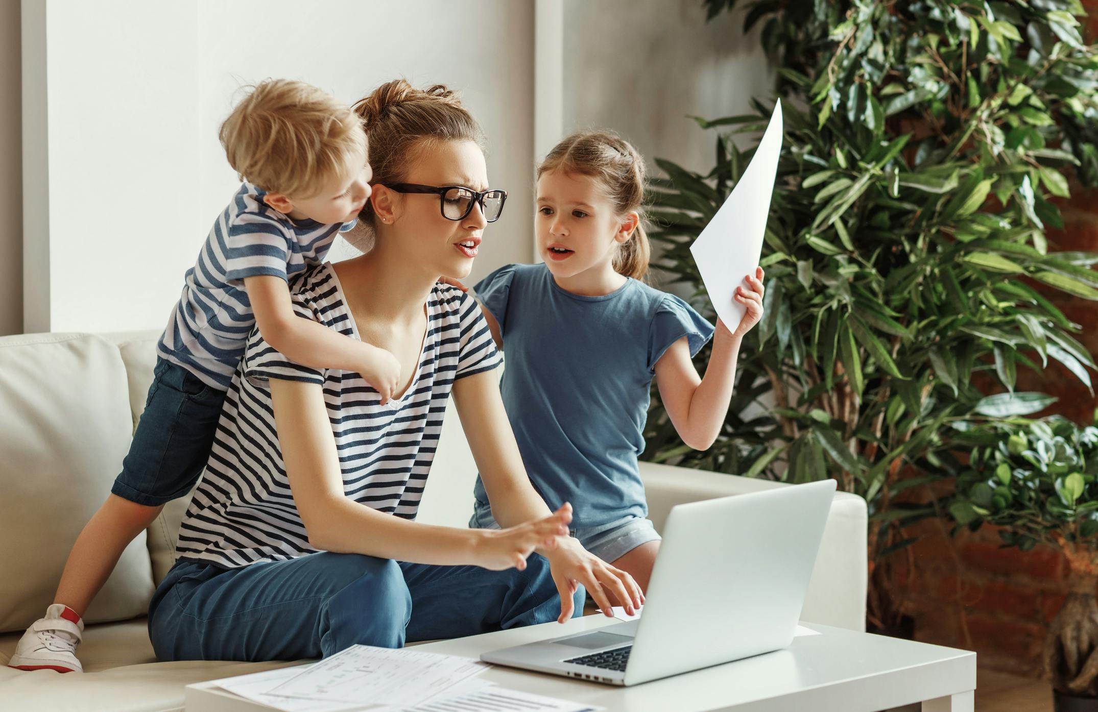 Busy mom on computer with her two kids behind her.