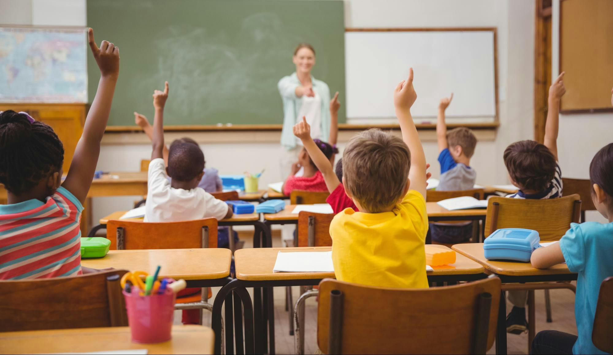 A teacher calls on a student wearing a yellow shirt while the entire class raises their hands to answer a question.