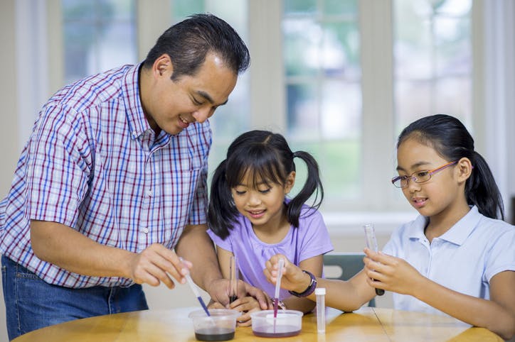 A father helps his two young daughters with a science experiment.