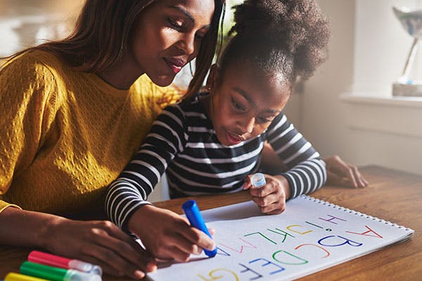 A monther is sitting beside her daughter, instructing her to draw letters