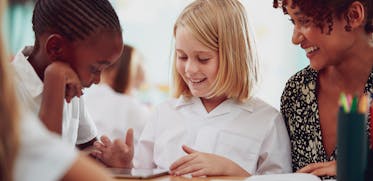 A teacher sitting with two elementary students as they learn in class.