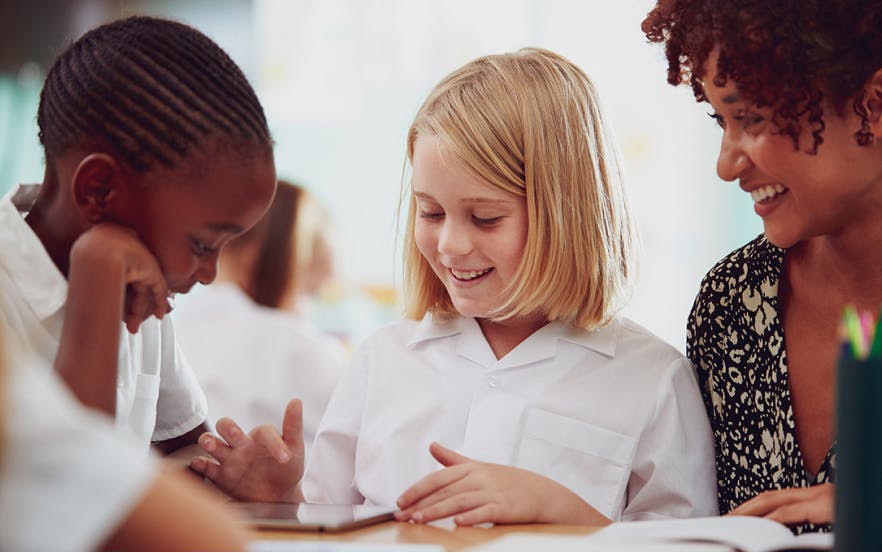 A teacher sitting with two elementary students as they learn in class.