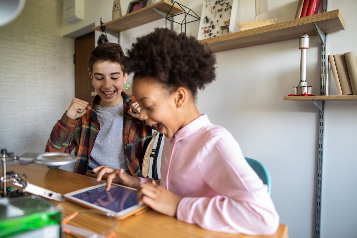 Two children sitting at a table, excitedly playing a game on a tablet device.