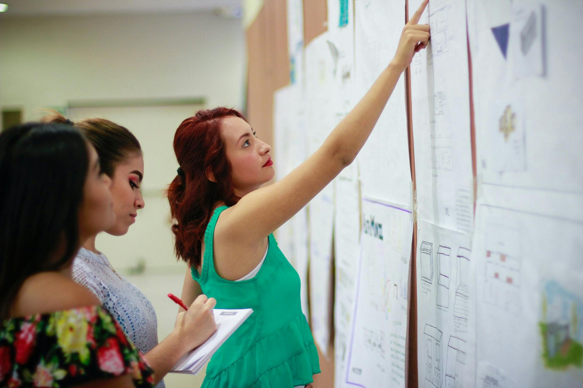 Three women stand in front of a board, one woman with red hair and a green shirt points at a chart while the other two take notes. 