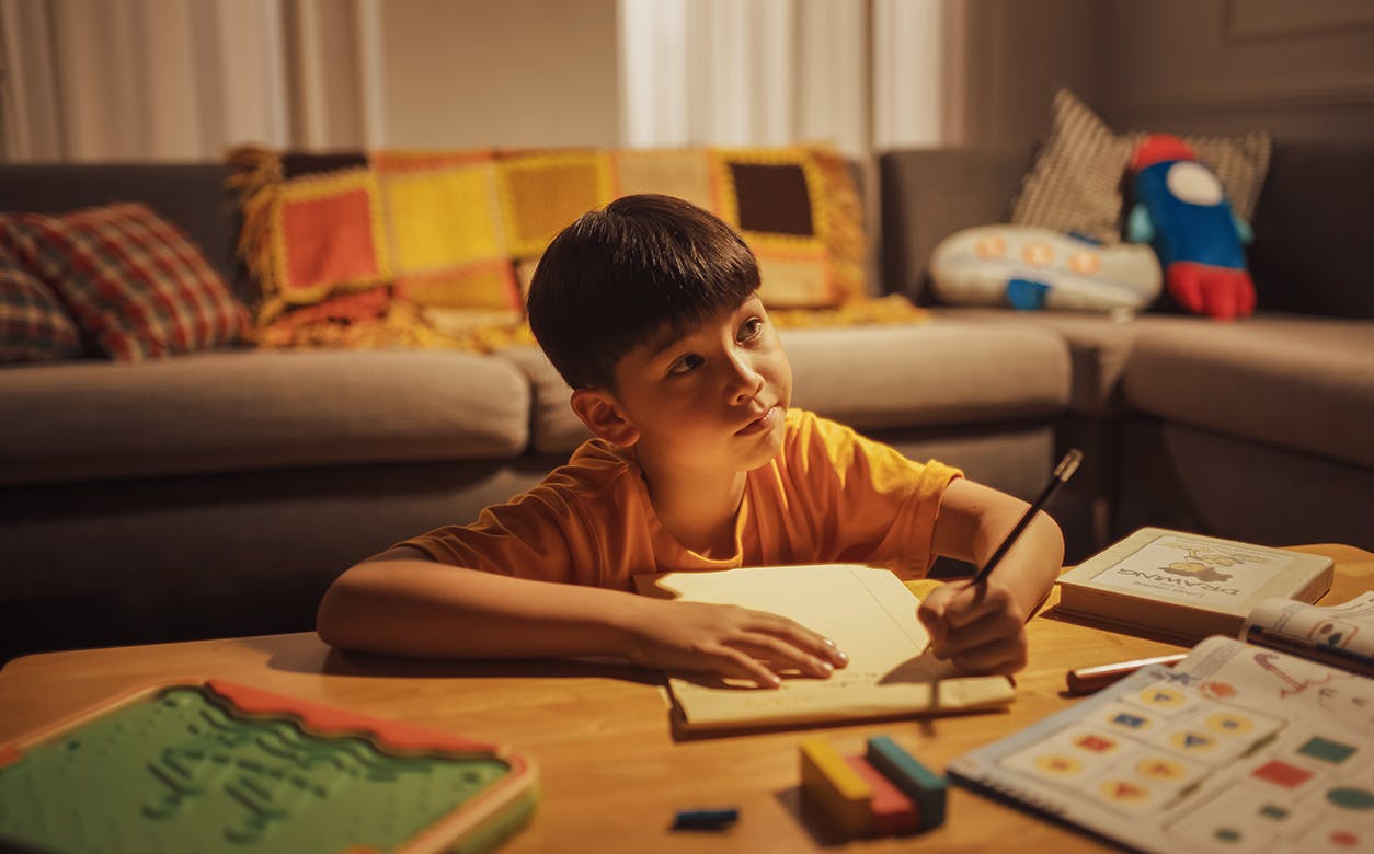 A child sits at a table, concentrating as he writes with a pen on a sheet of paper